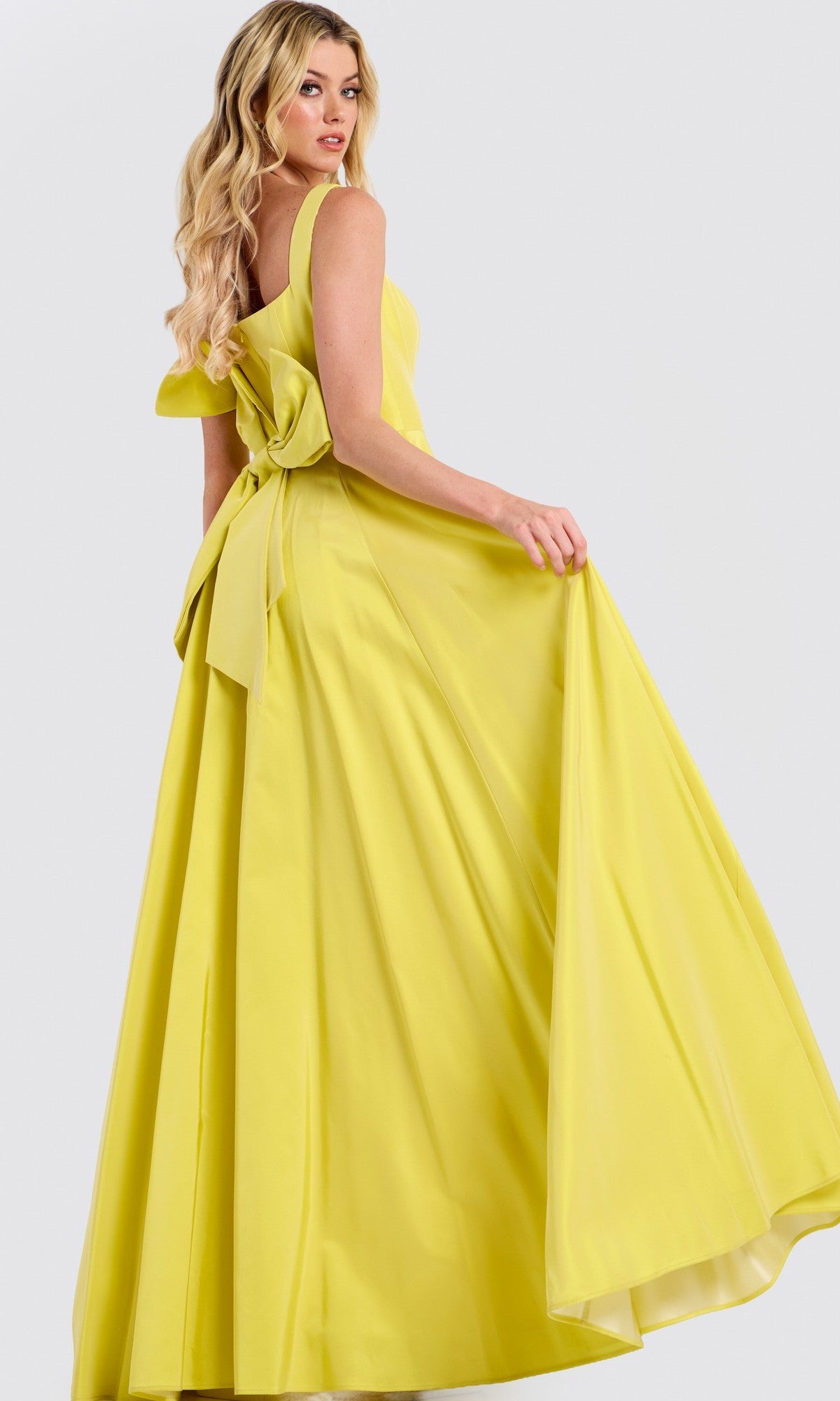 A woman models the Jovani 47496 long prom dress, featuring a bright yellow hue, an a-line flowy skirt, and a large back bow. She stands sideways, holding out one side of her skirt.